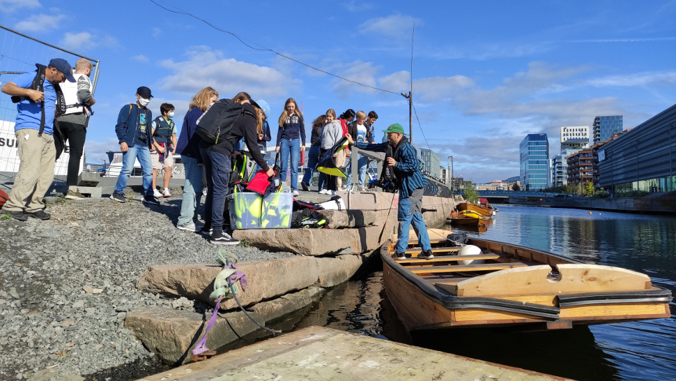 Elever står ved elvebredden og venter på å gå om bord i en båt